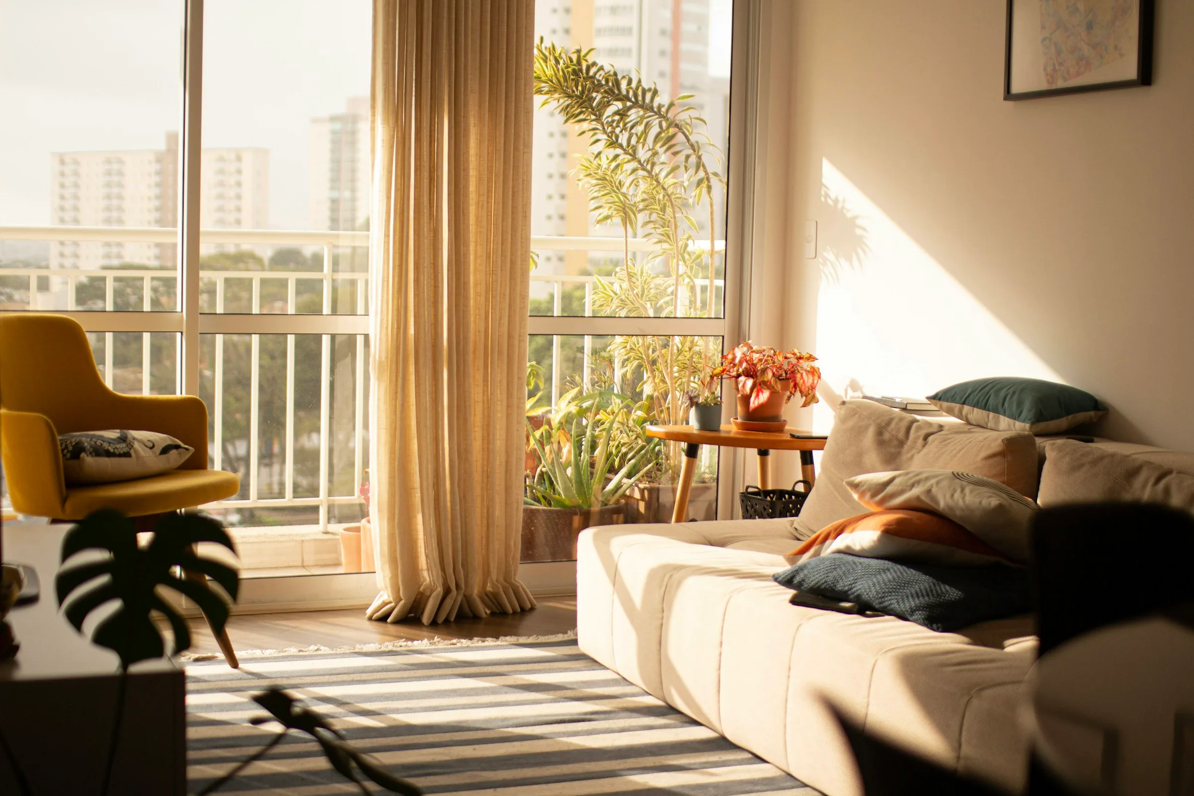A sunlit living room with a cream sofa, a mustard armchair and tall windows with long curtains