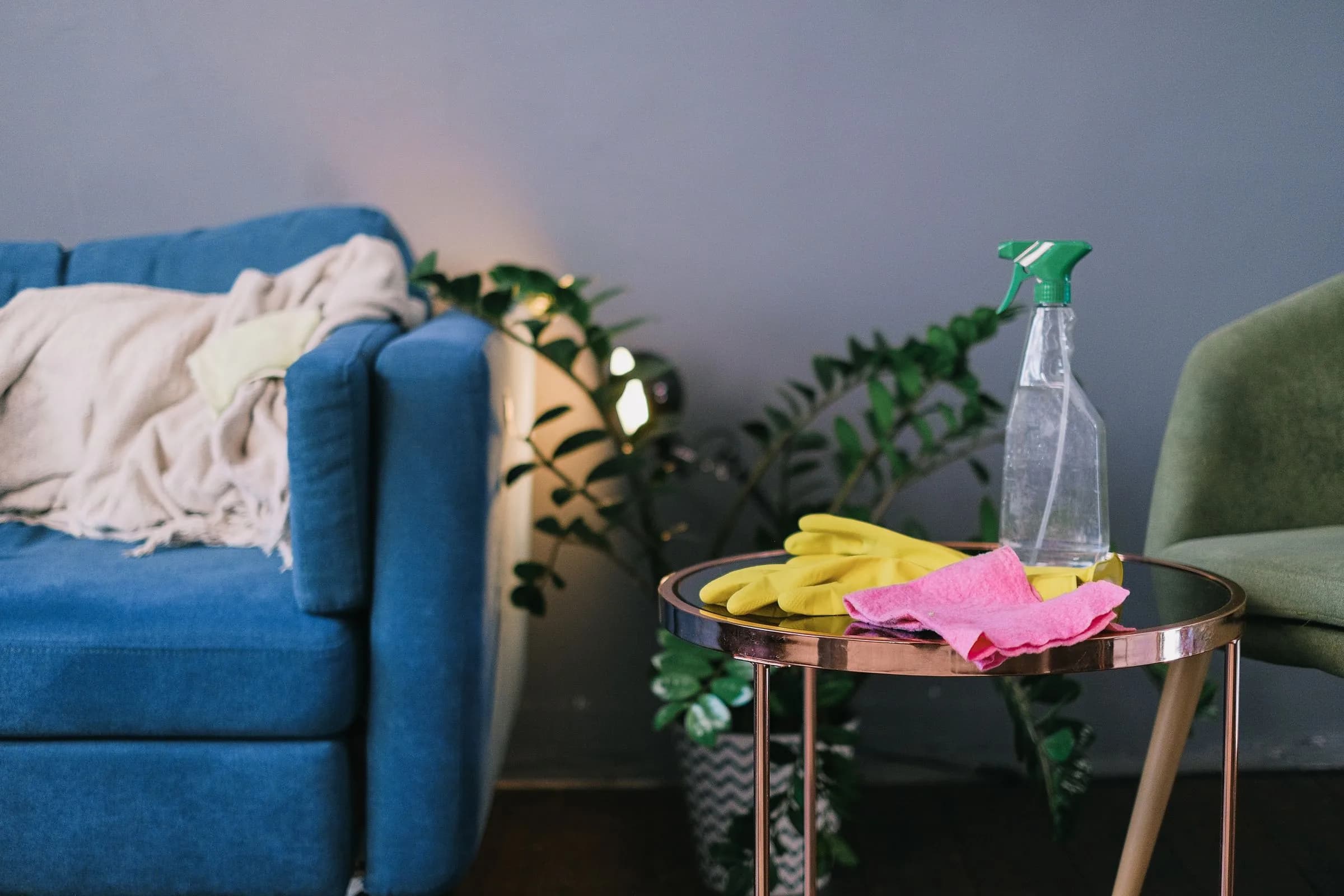A spray bottle, yellow rubber gloves and a pink cloth on a small round table beside a blue sofa and a pot plant.