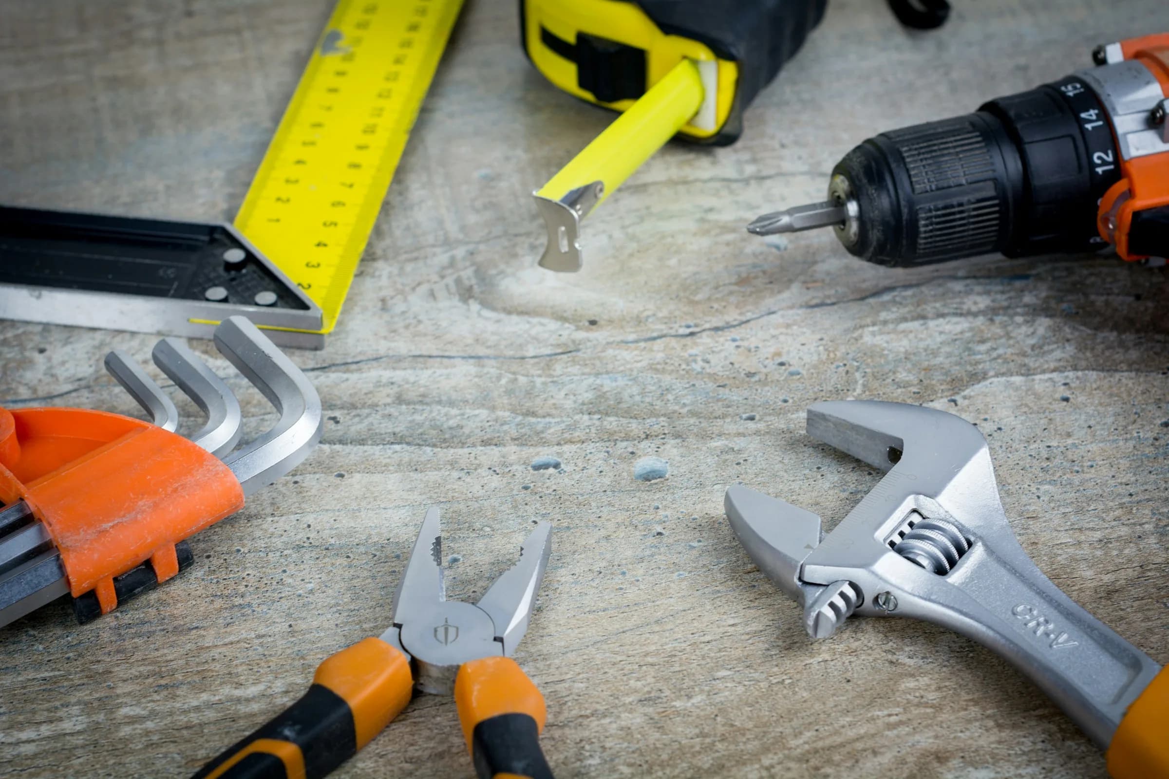 A spread of hand tools, including a tape measure, drill, pliers and adjustable wrenches, on a worn table