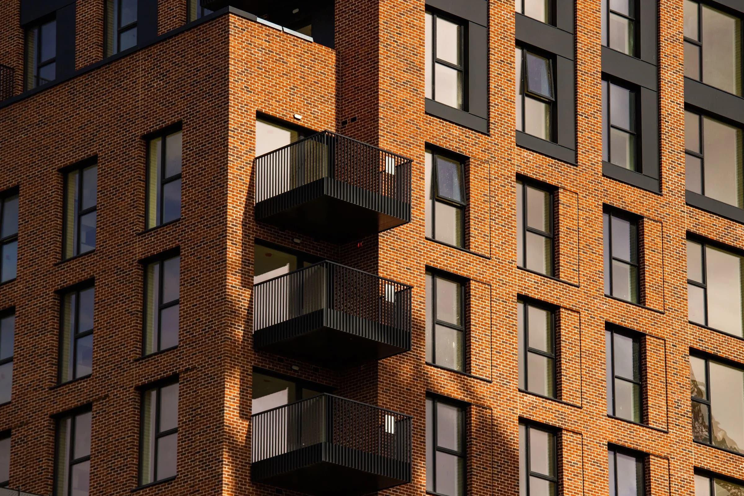A modern red-brick apartment building with dark-framed windows and balconies in sunlight.