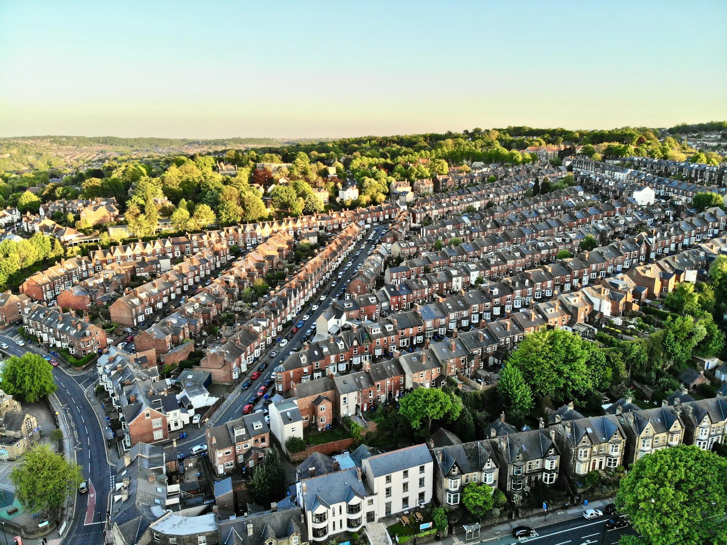 An aerial view of long rows of terraced houses and tree-lined streets in warm evening light.