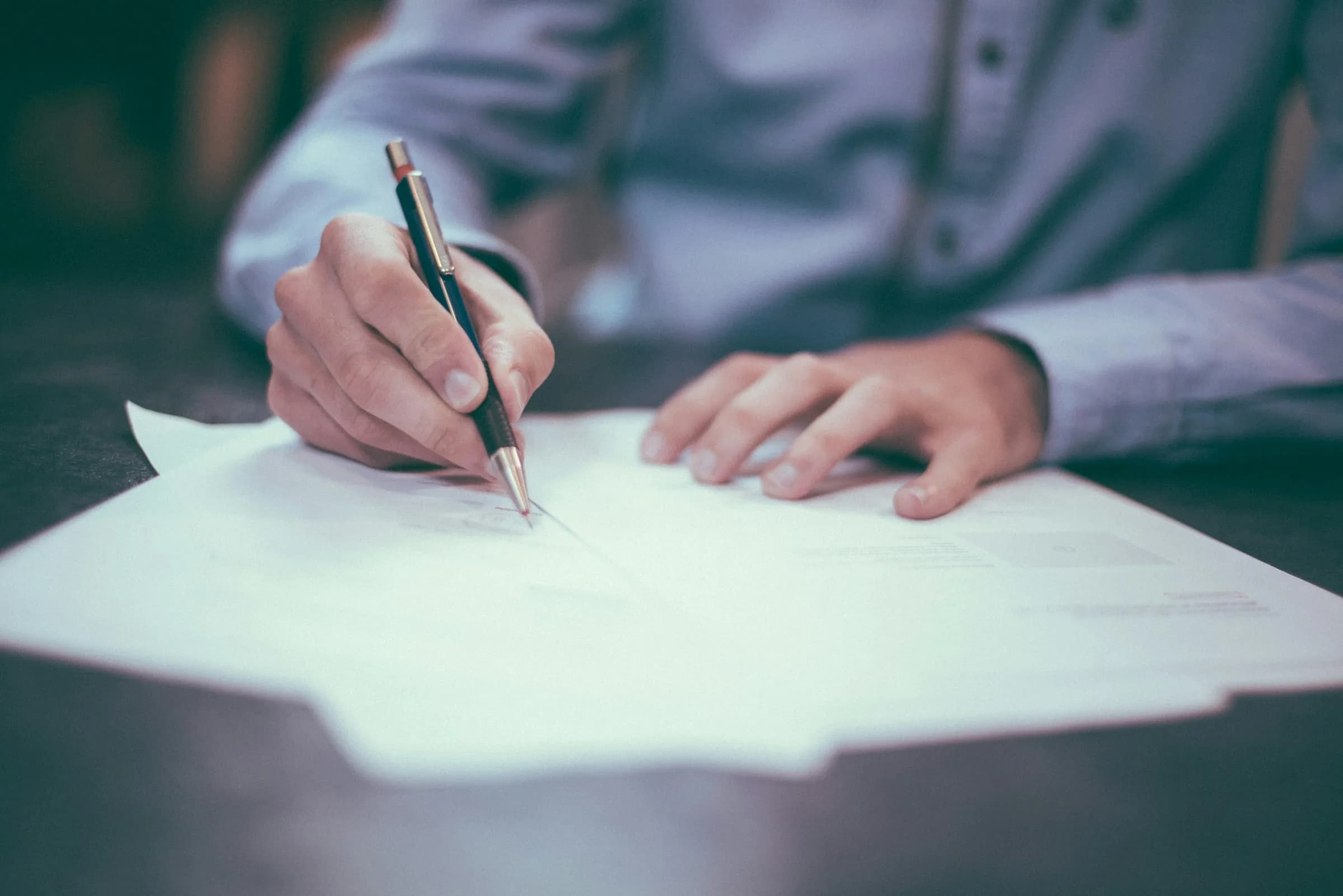 A person in a blue shirt signing a document with a pen at a dark table, face out of frame.