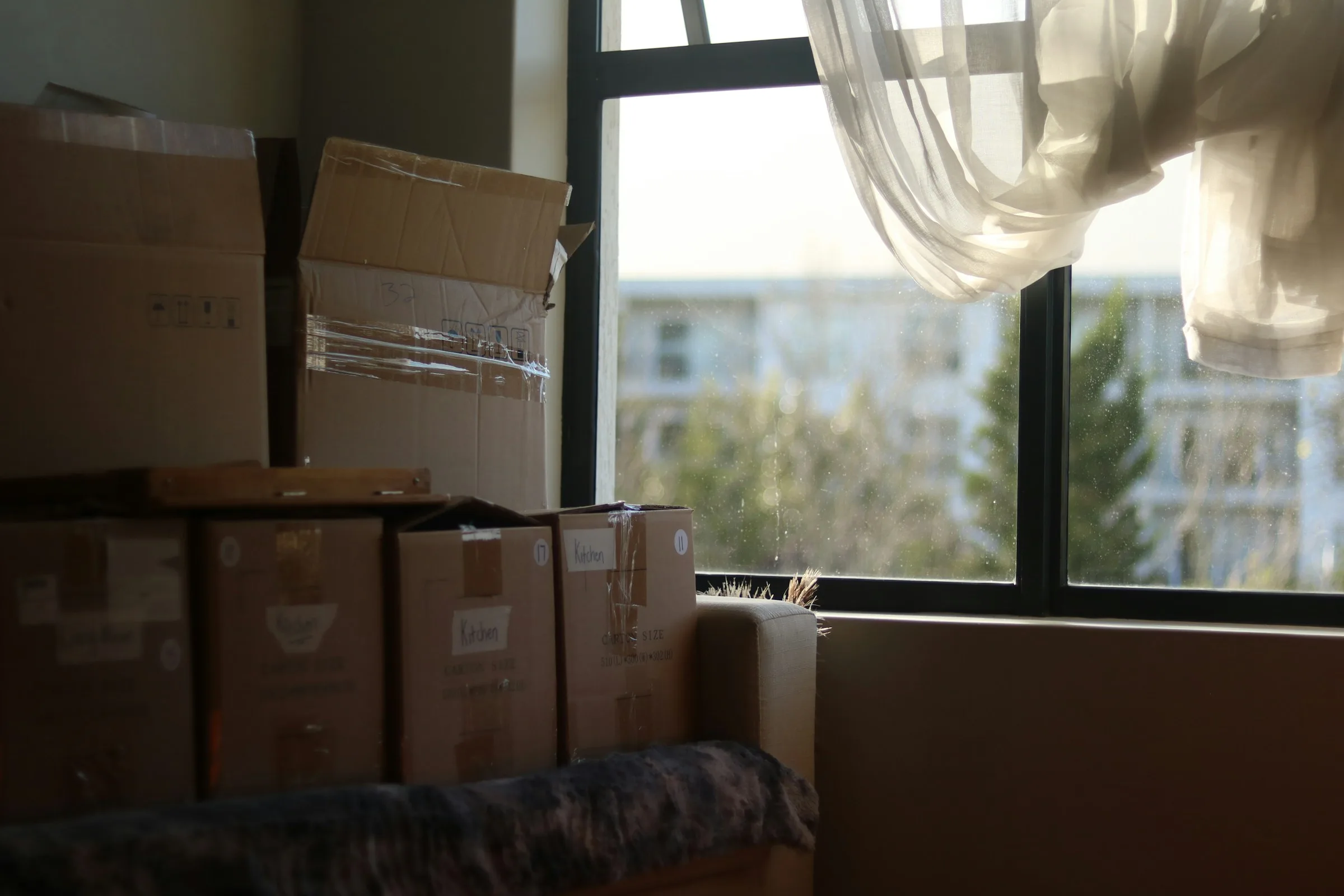 Sealed cardboard boxes labelled Kitchen stacked beside a window with a white curtain in a softly lit room.