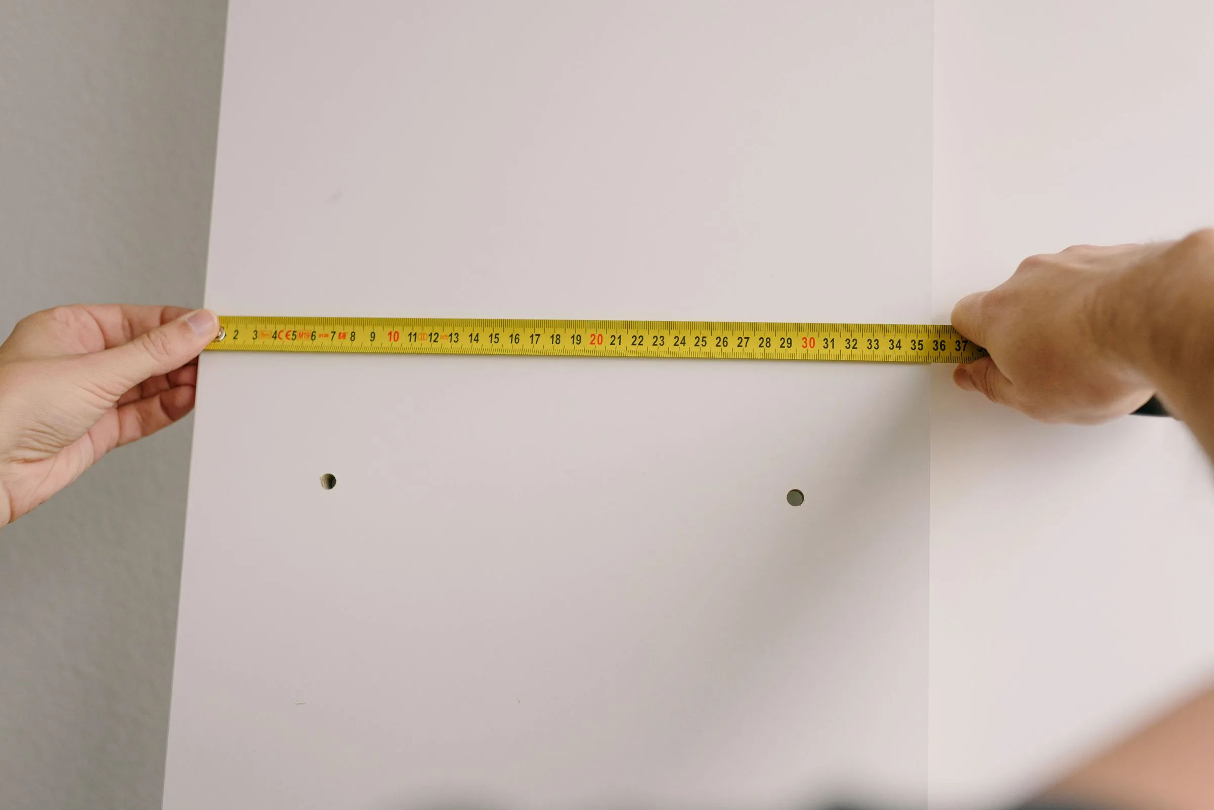 Two hands holding a yellow tape measure flat against a white wall.