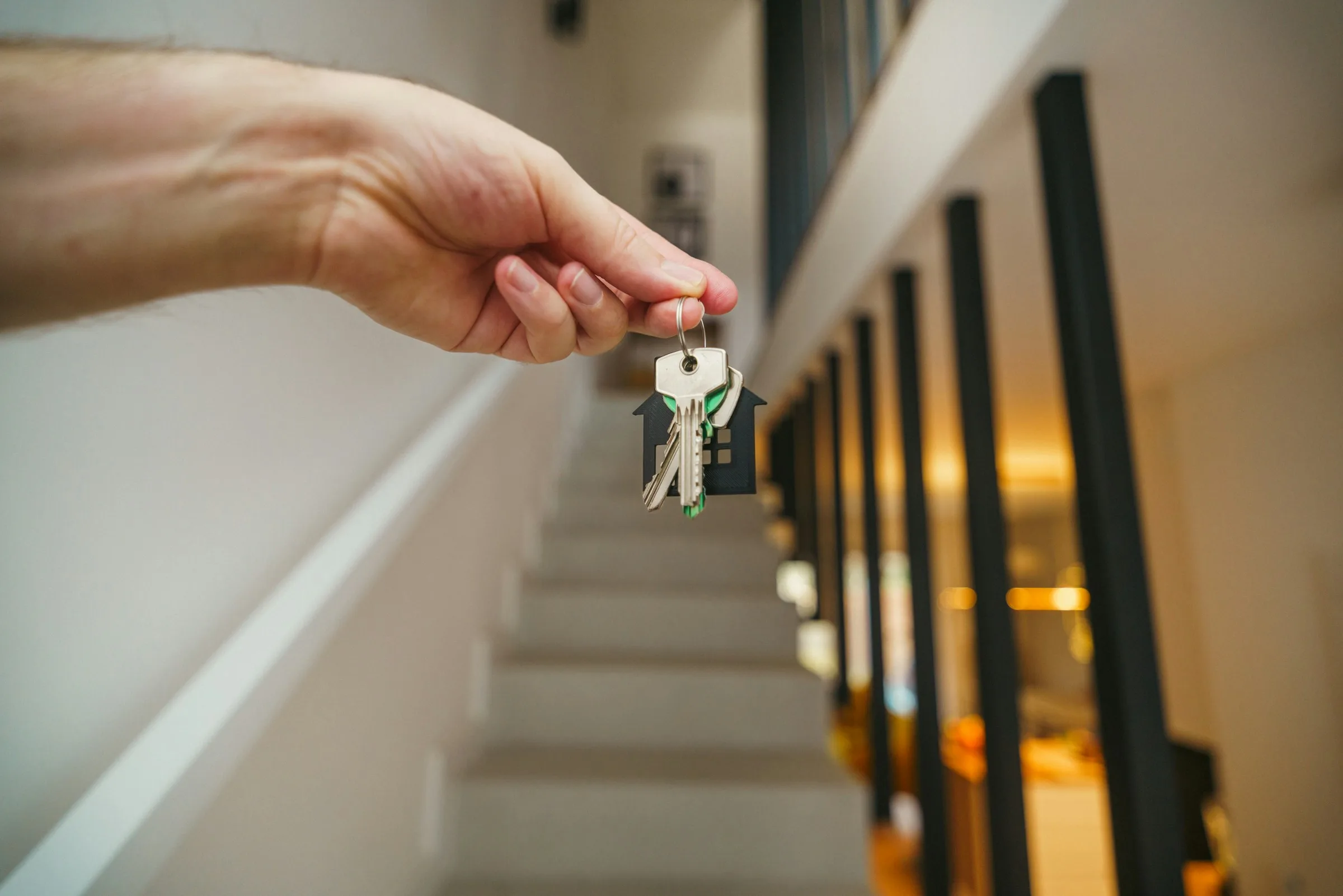 A hand holding a set of keys on a small house-shaped keyring above a staircase.