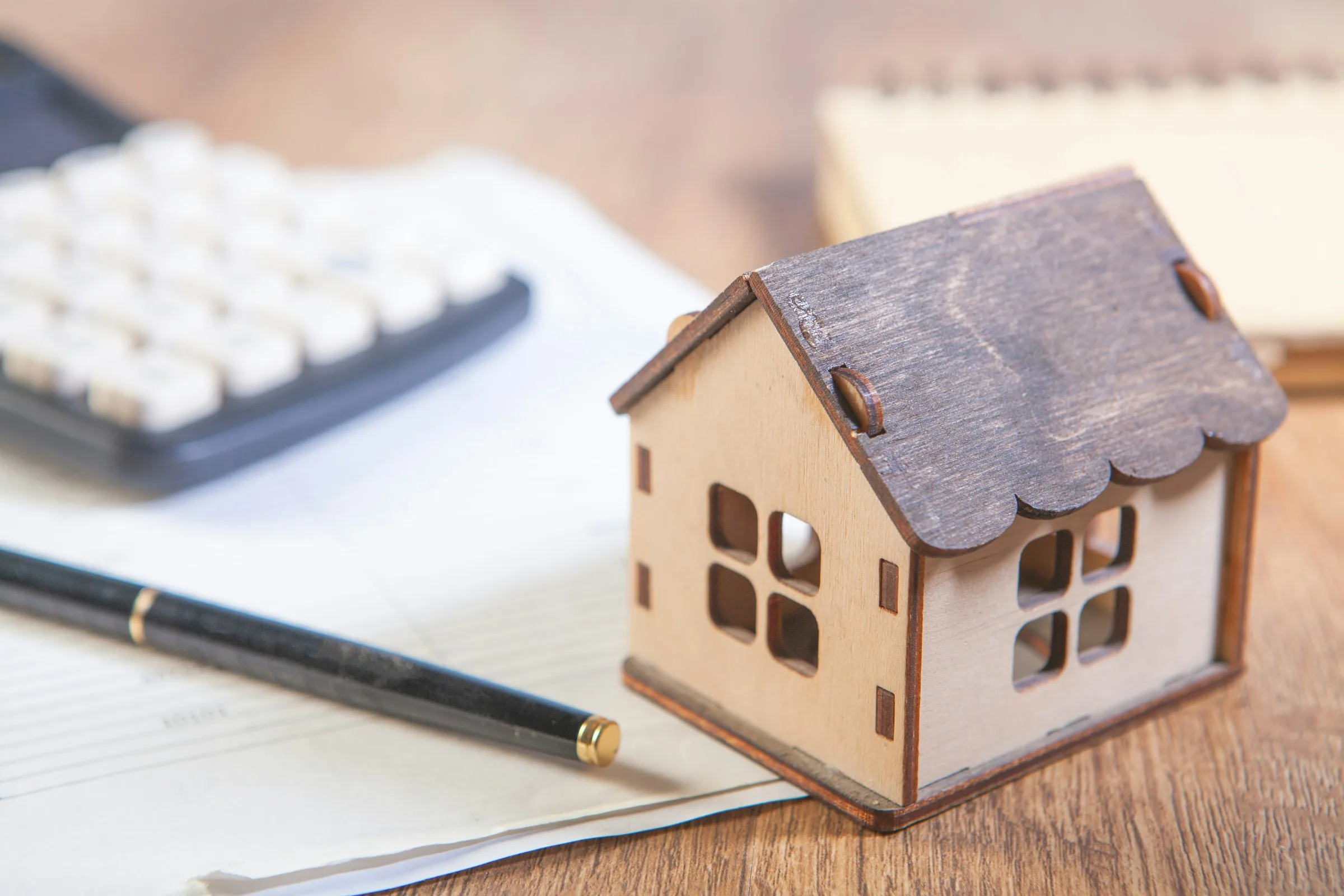 A small wooden model house beside a calculator, a pen and a notepad on a desk