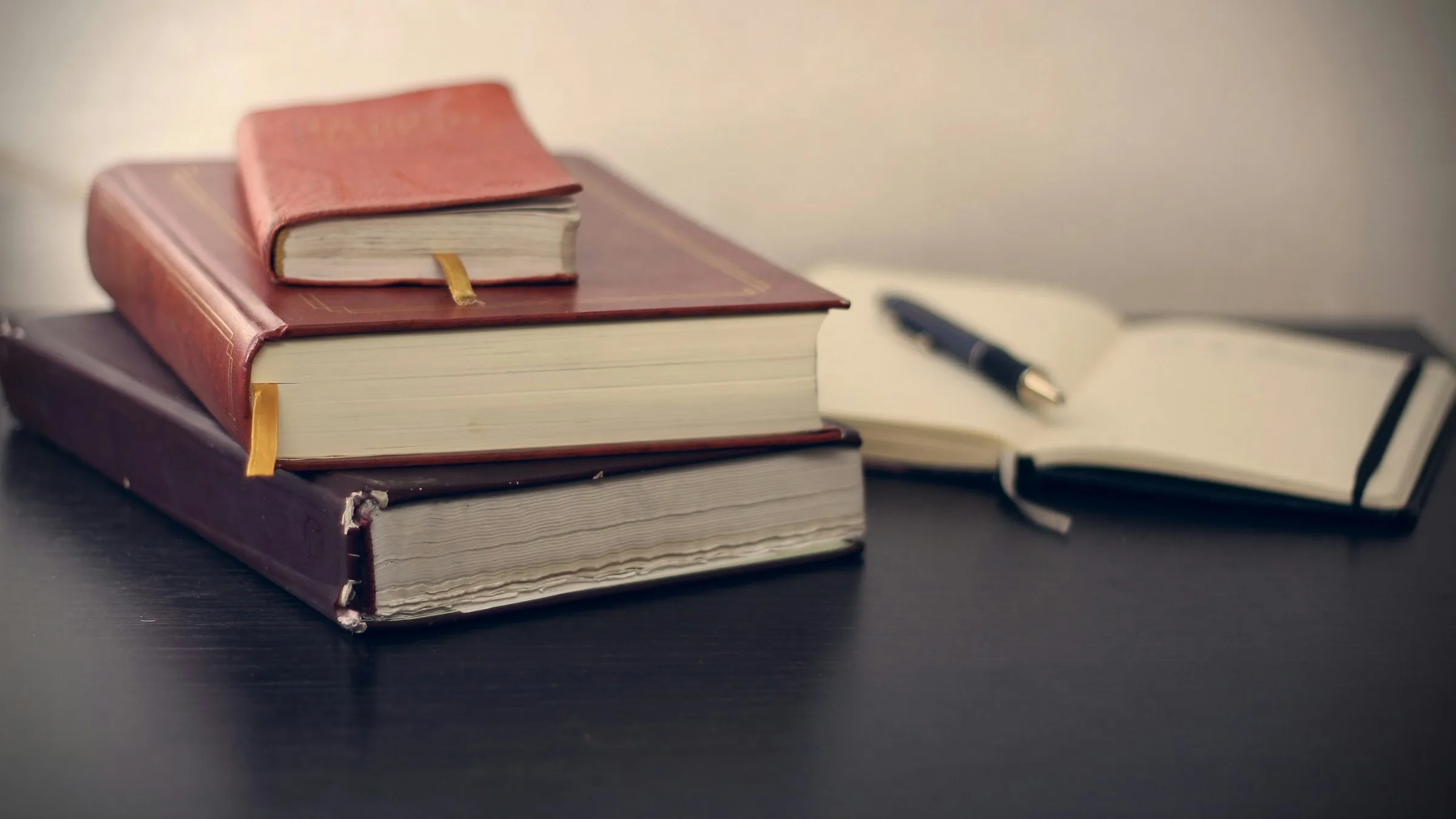 A stack of three hardback books beside an open notebook and pen on a dark table