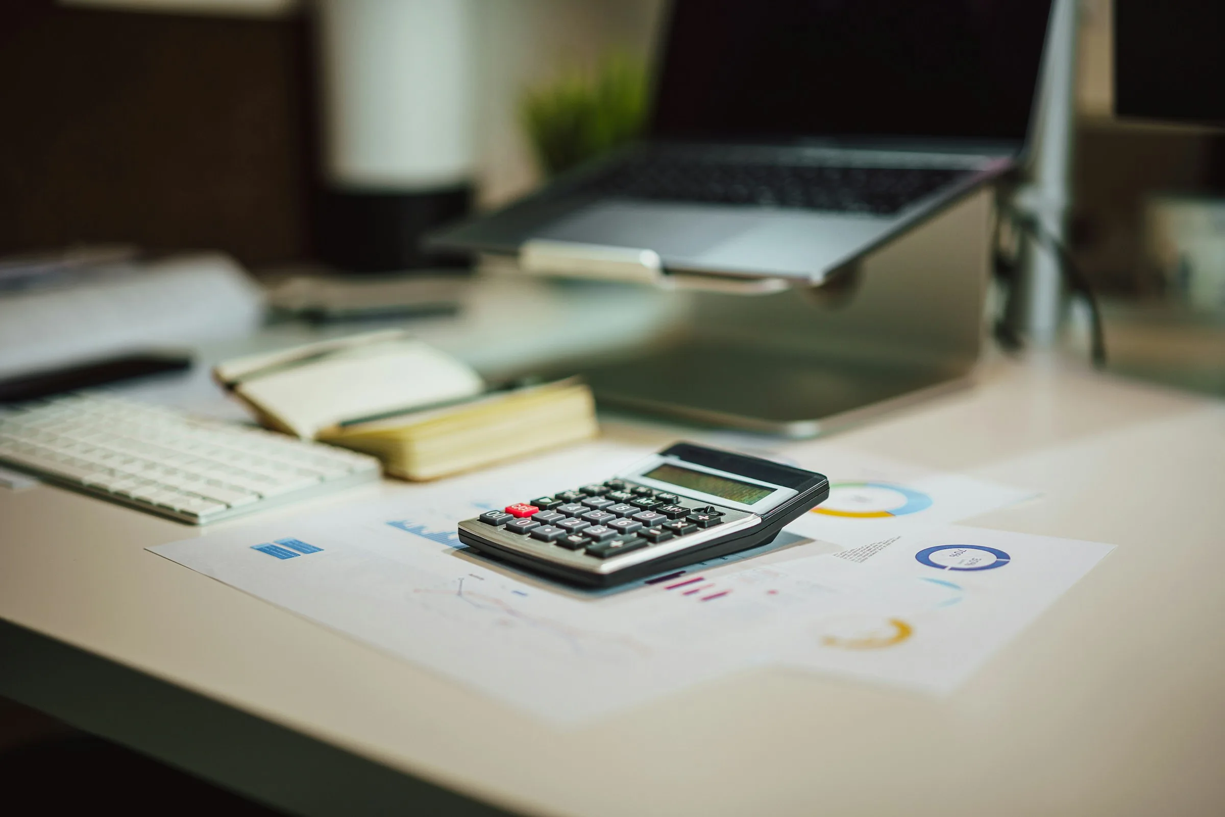 A desk calculator on printed charts beside a laptop and notepads, seen from above.