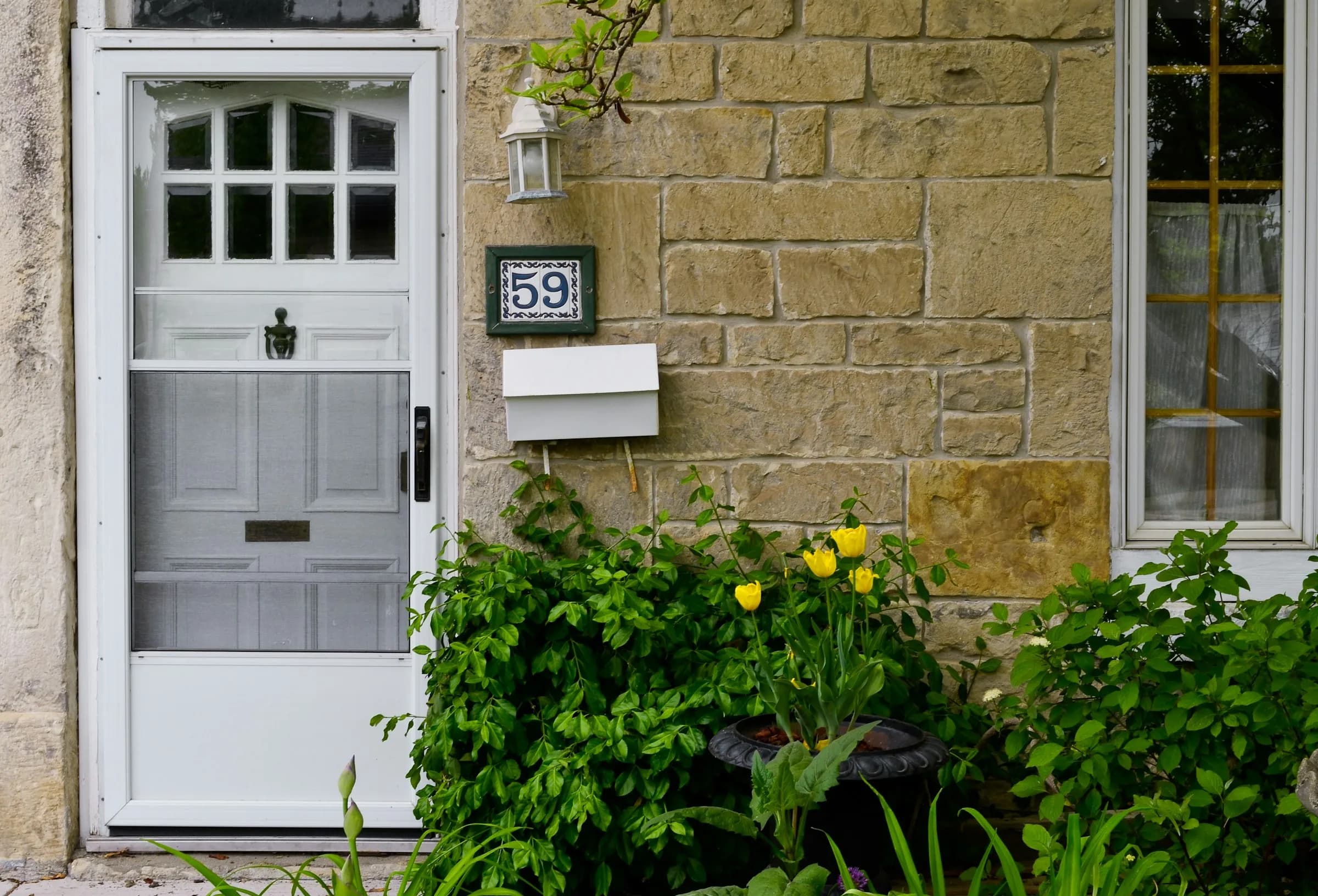 A white panelled front door set in a honey-coloured stone wall, framed by green plants and yellow flowers