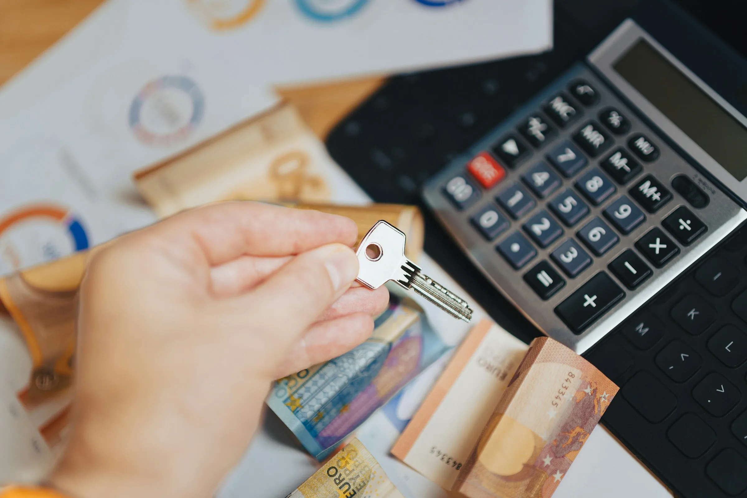 A hand holding a house key over a calculator, banknotes and printed charts