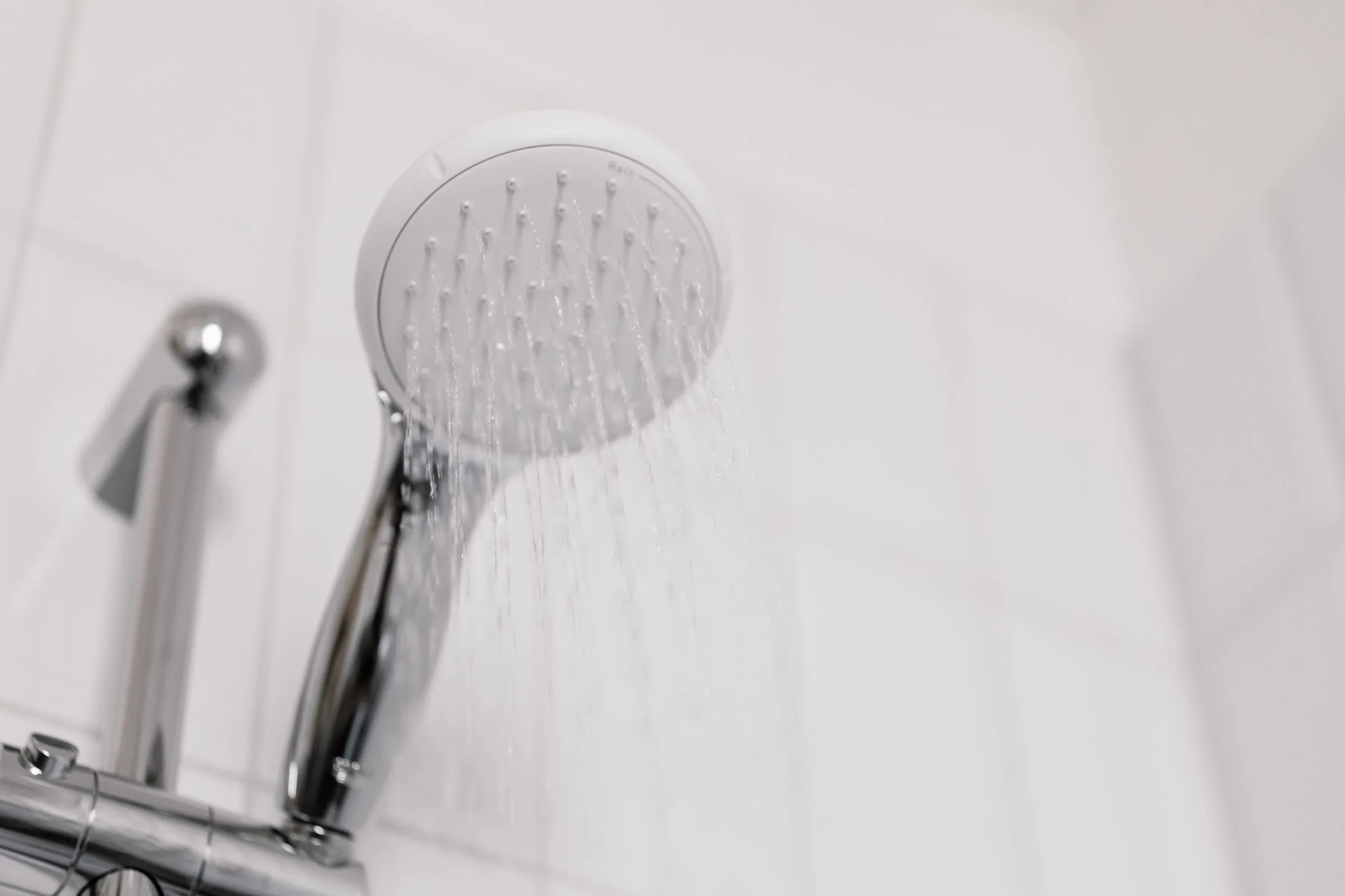 A white handheld shower head spraying water against white bathroom tiles beside a chrome fitting.