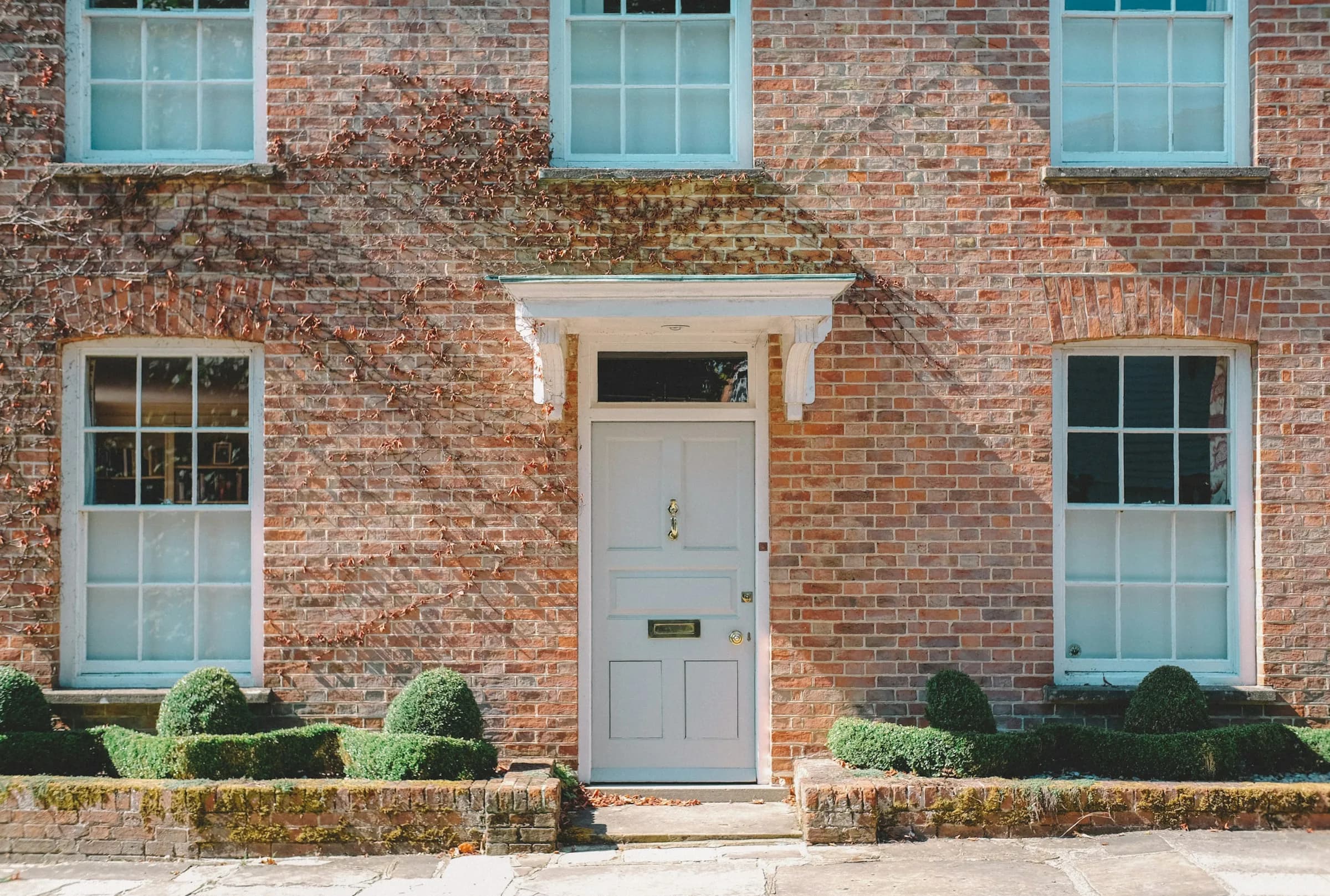 A red-brick house with sash windows and a white front door behind clipped hedges