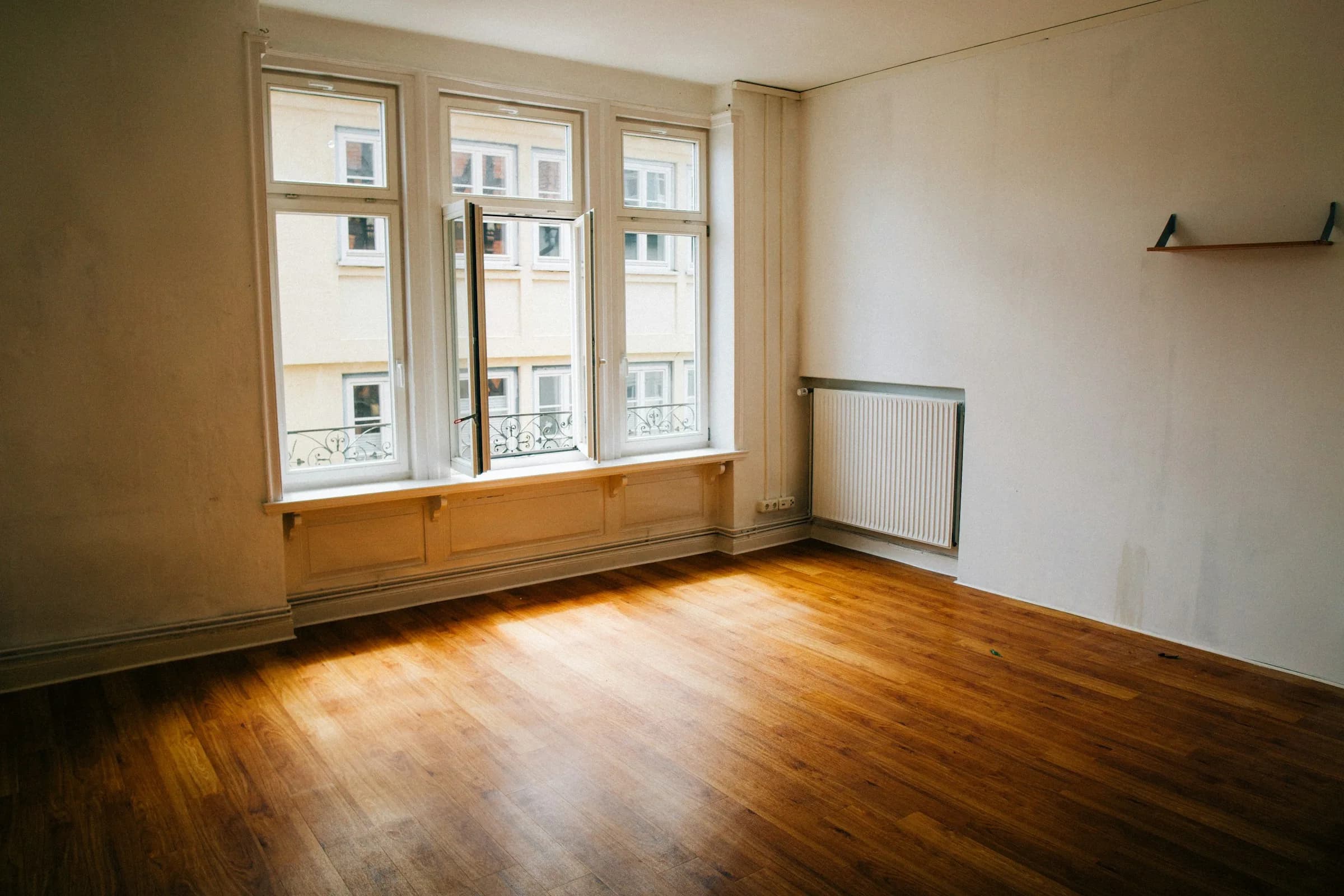 An empty room with a wooden floor, a tall window open onto a street, and a white radiator on the wall.