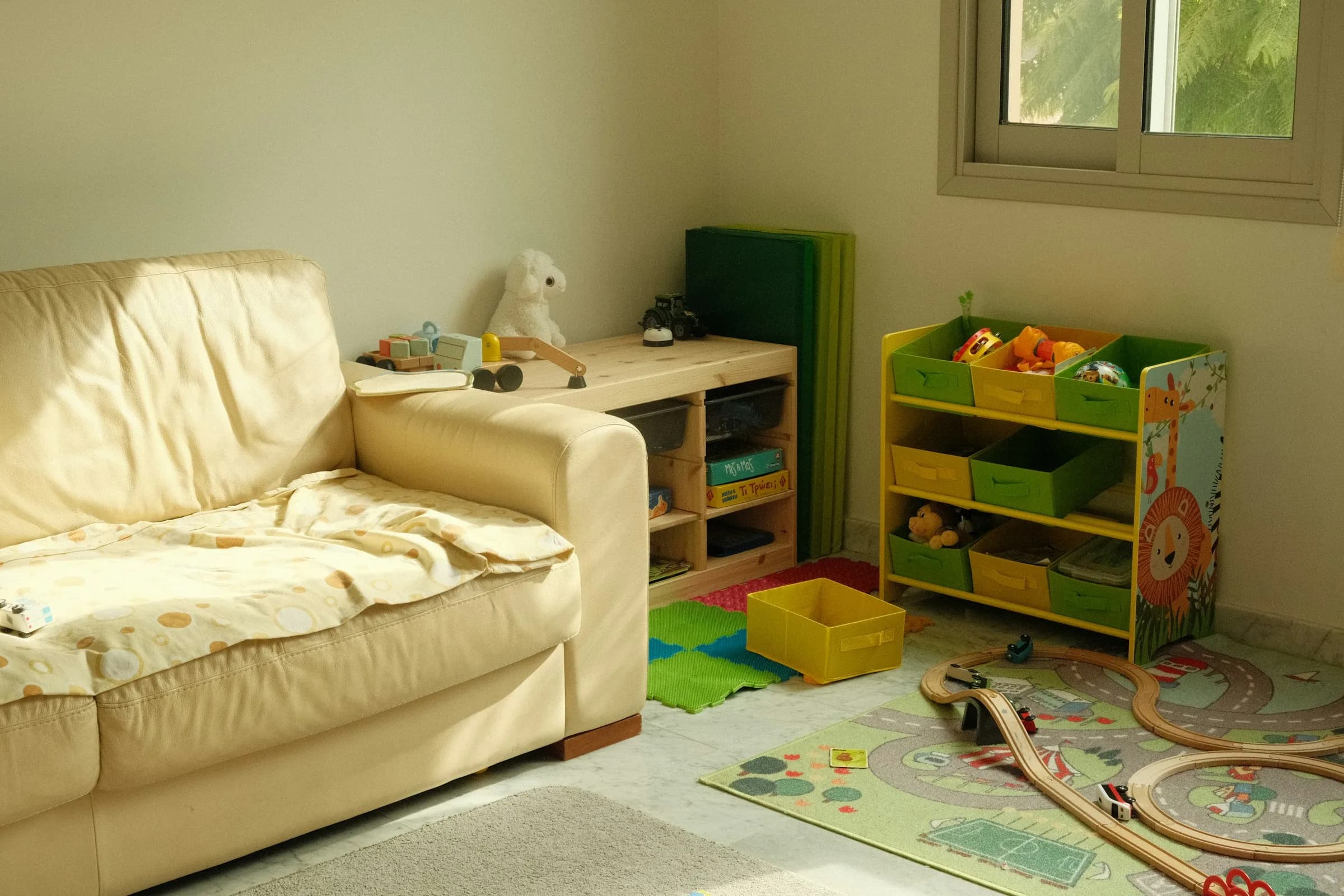 A bright corner of a living room with a cream sofa, a toy storage shelf and a wooden train set on a play mat.