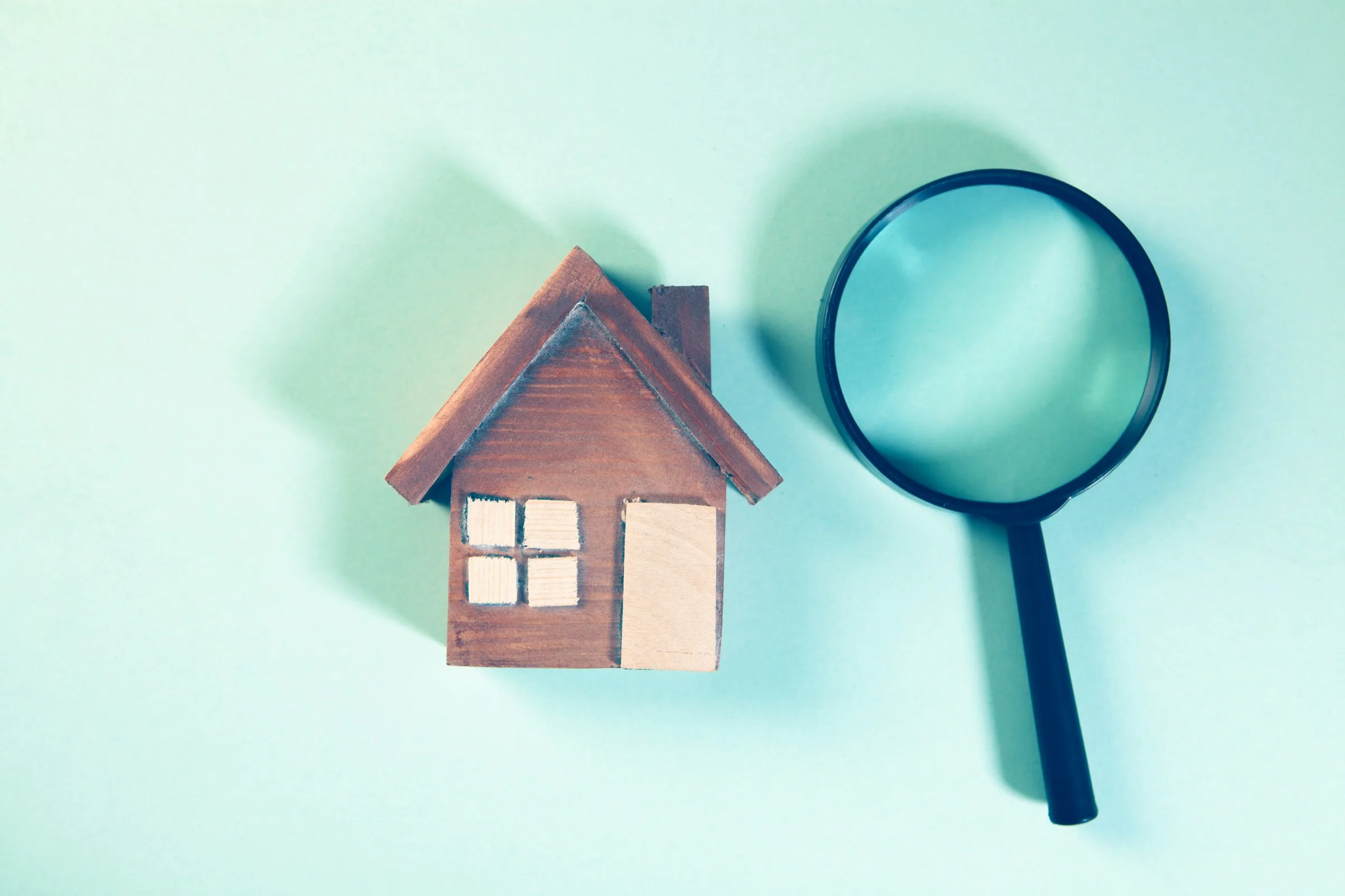 A magnifying glass beside a small wooden model house on a pale green background