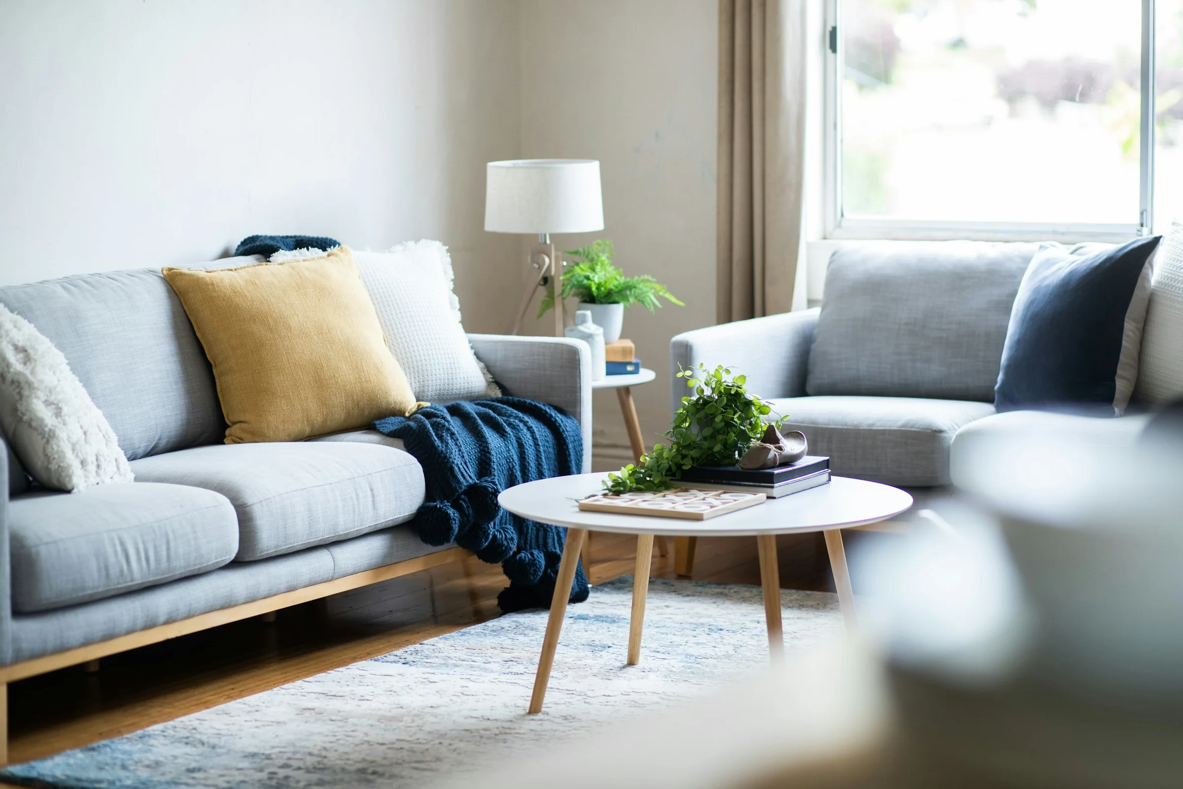 A grey fabric sofa with mustard and white cushions and a navy knitted throw beside a round white coffee table and a lamp in a bright living room.