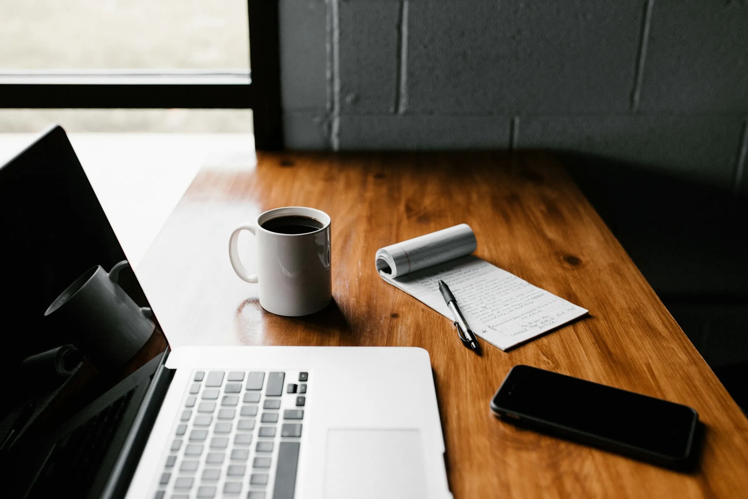 An open laptop, a mug of coffee, a notepad with a pen and a phone on a wooden desk beside a window.