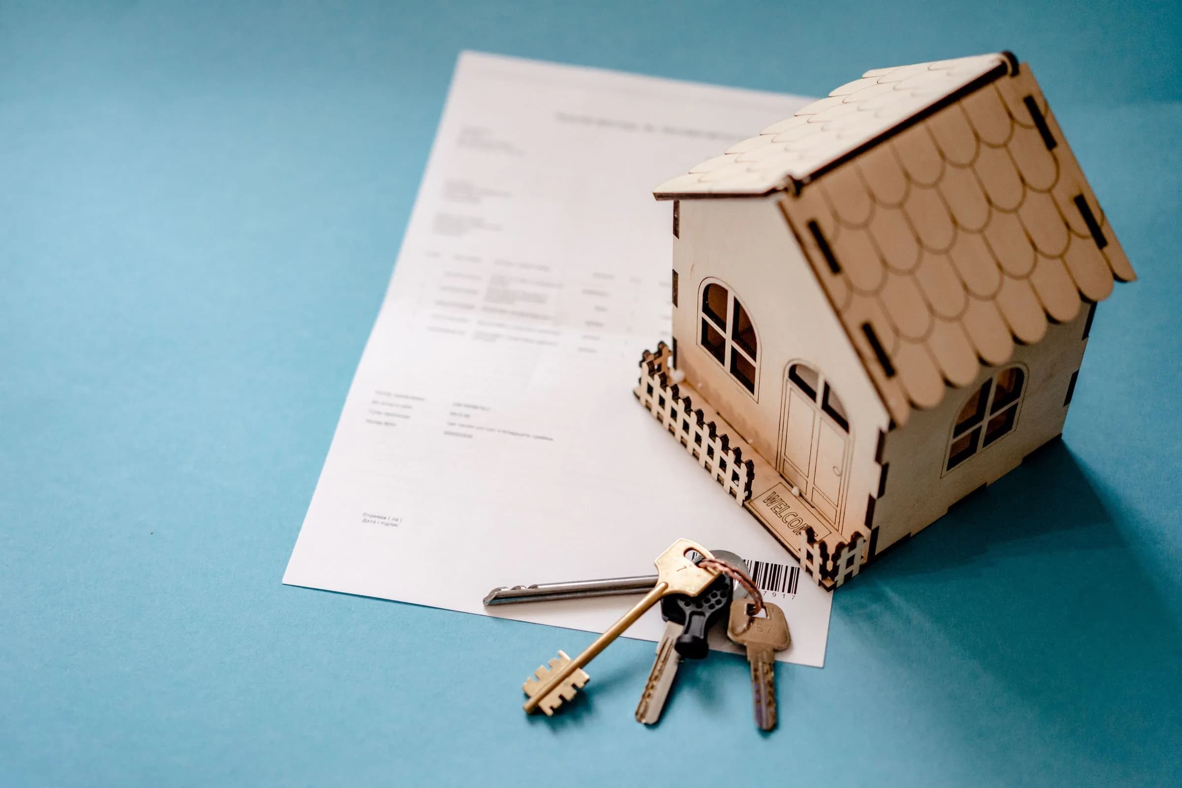 A small wooden model house and a set of keys resting on a printed contract on a blue table.
