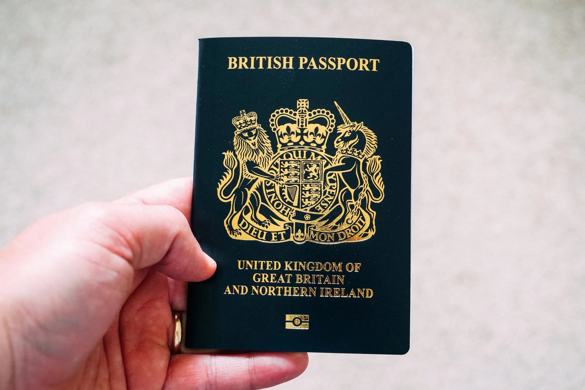 A hand holding a British passport with its gold crest on a dark green cover, against a pale grey background.