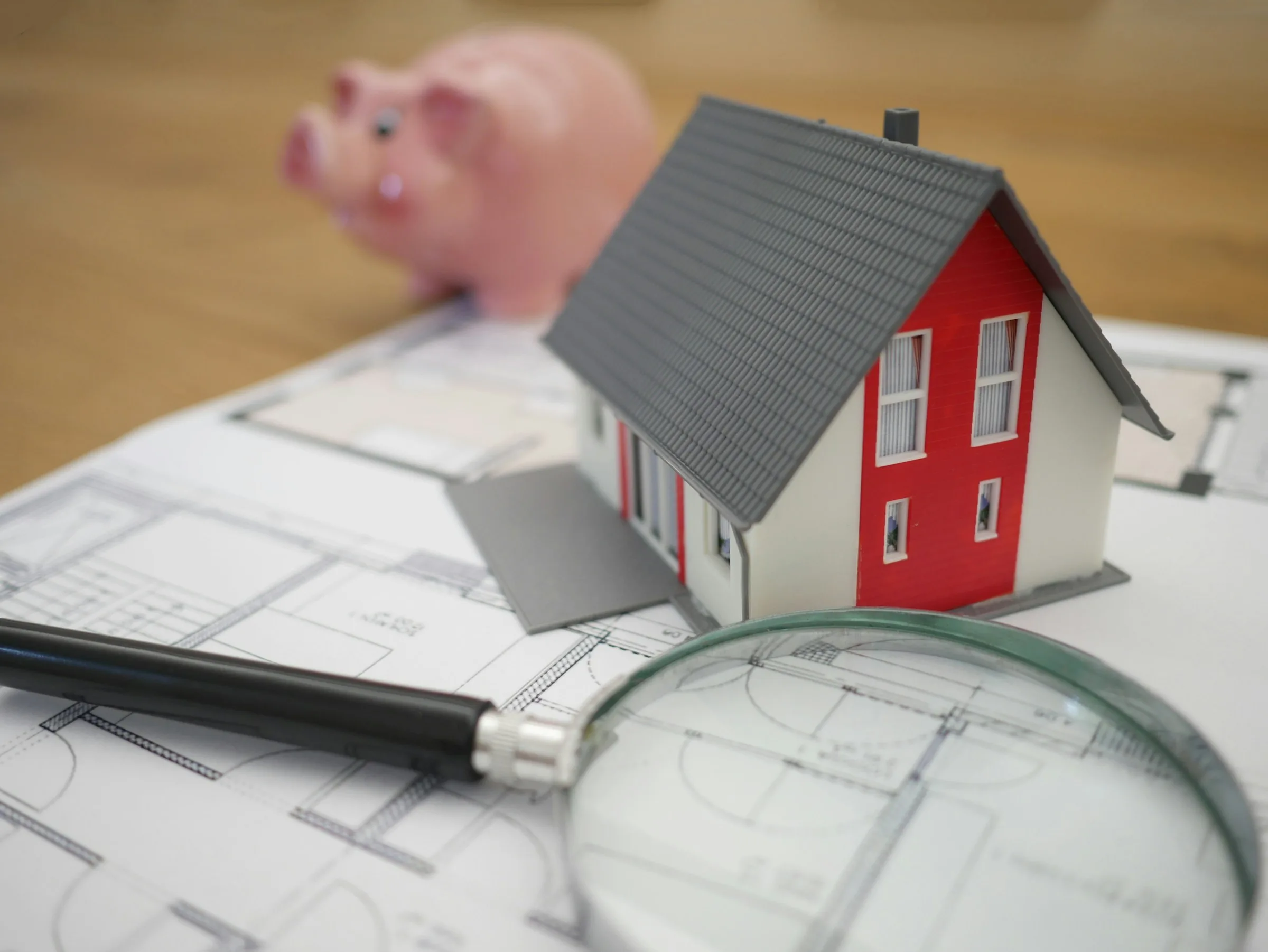 A small white and red model house on a printed floor plan, with a magnifying glass lying across it and a piggy bank behind