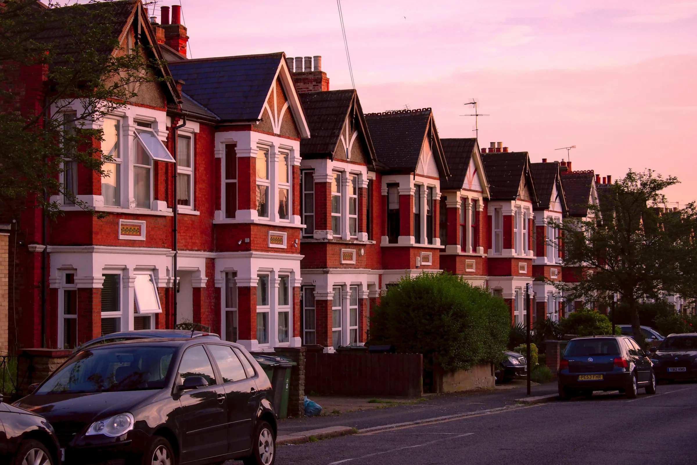 A terrace of red-brick houses with bay windows and pitched gables in warm evening light, with cars parked in front.