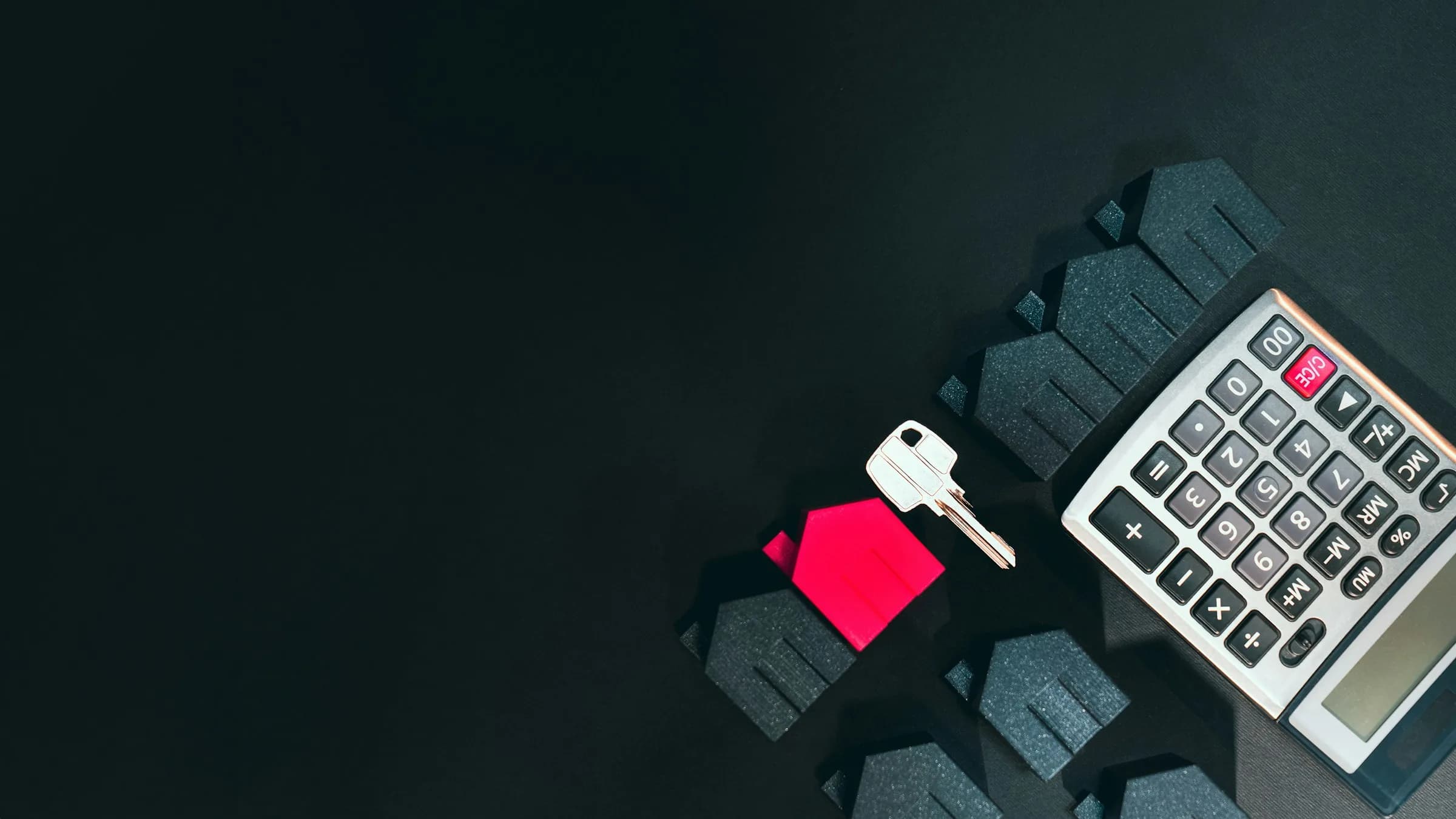 A house key and a calculator beside miniature grey and red model houses on a dark background.