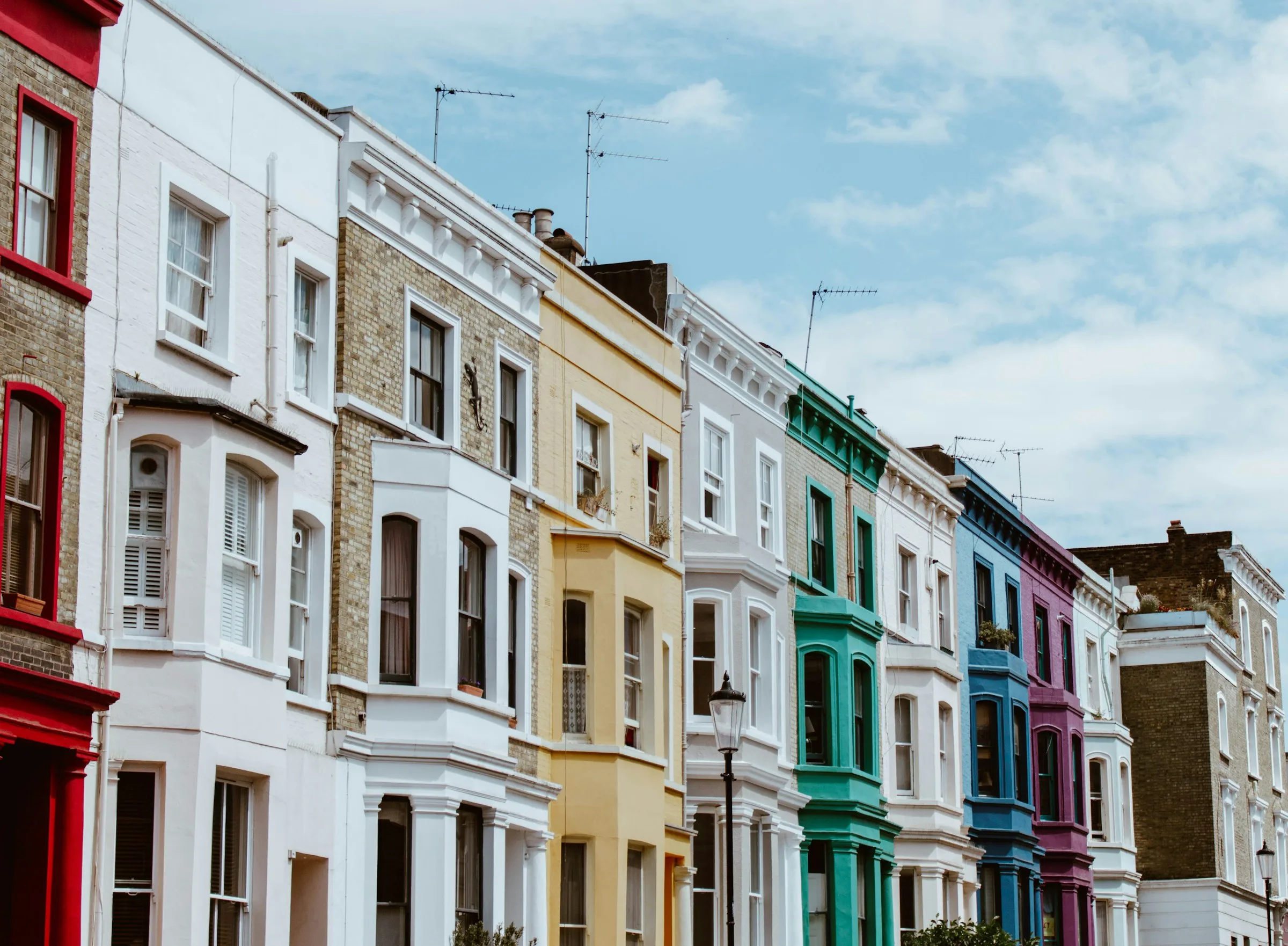 A row of terraced houses with bay windows painted white, yellow, green, blue and purple under a cloudy blue sky.
