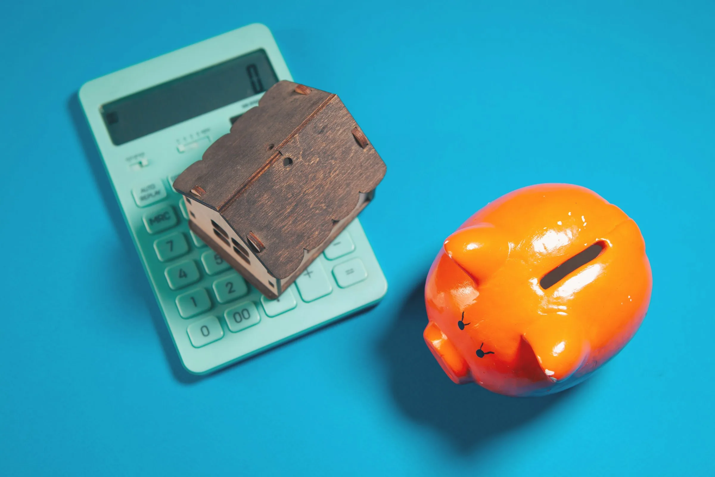 A mint-green calculator with a small wooden house model and an orange piggy bank on a blue background