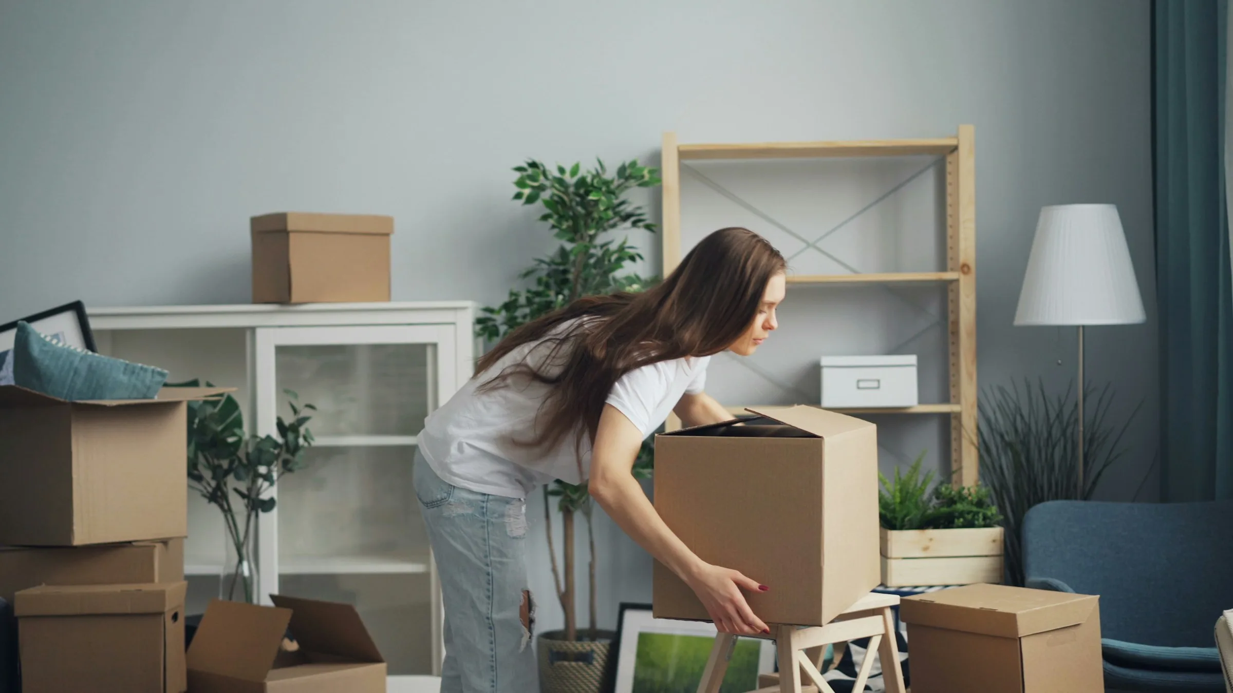 A woman lifting a cardboard box in a living room with shelves and houseplants