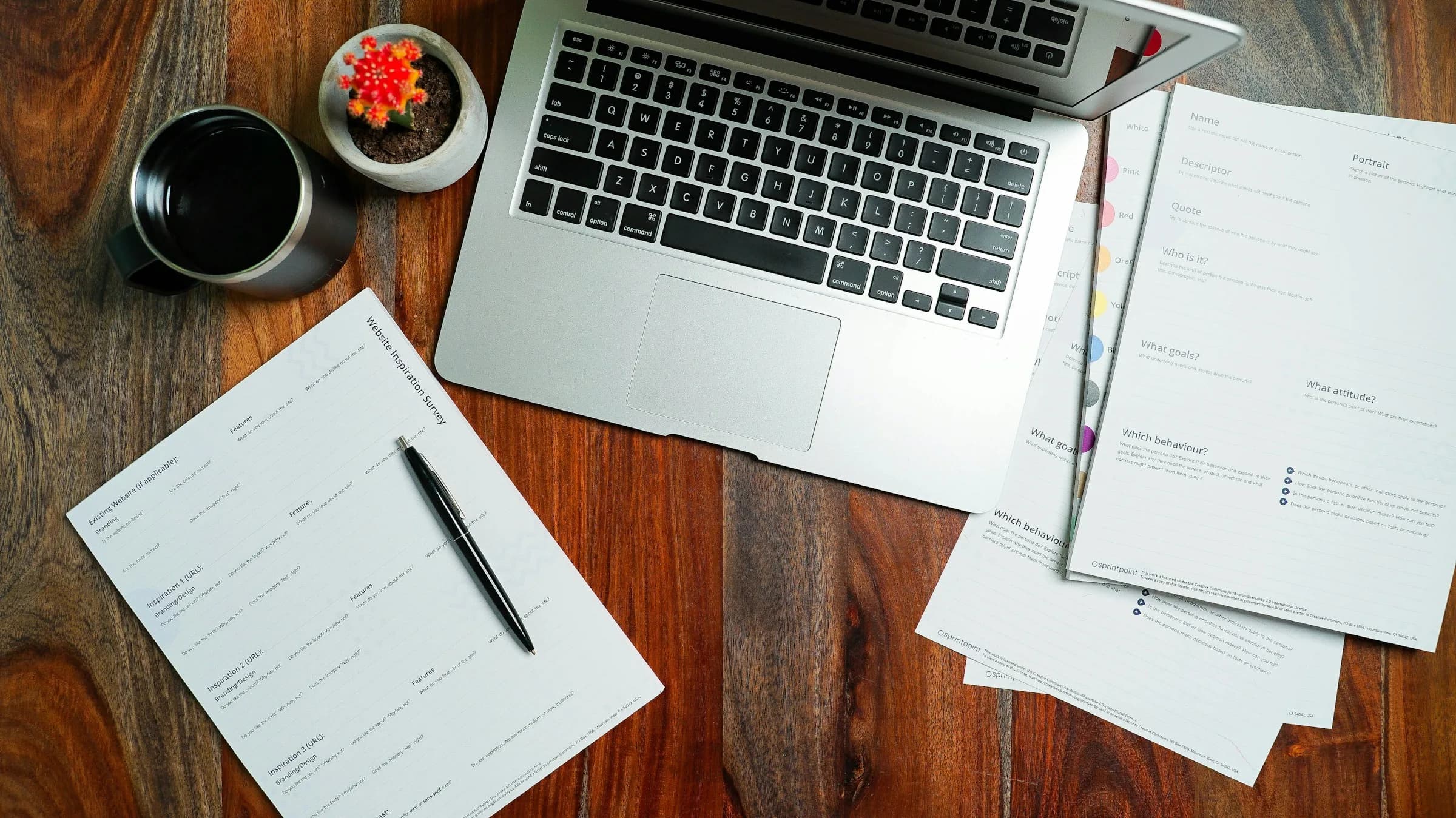 A laptop, a coffee cup, a pen and printed forms laid out on a wooden desk, seen from above.
