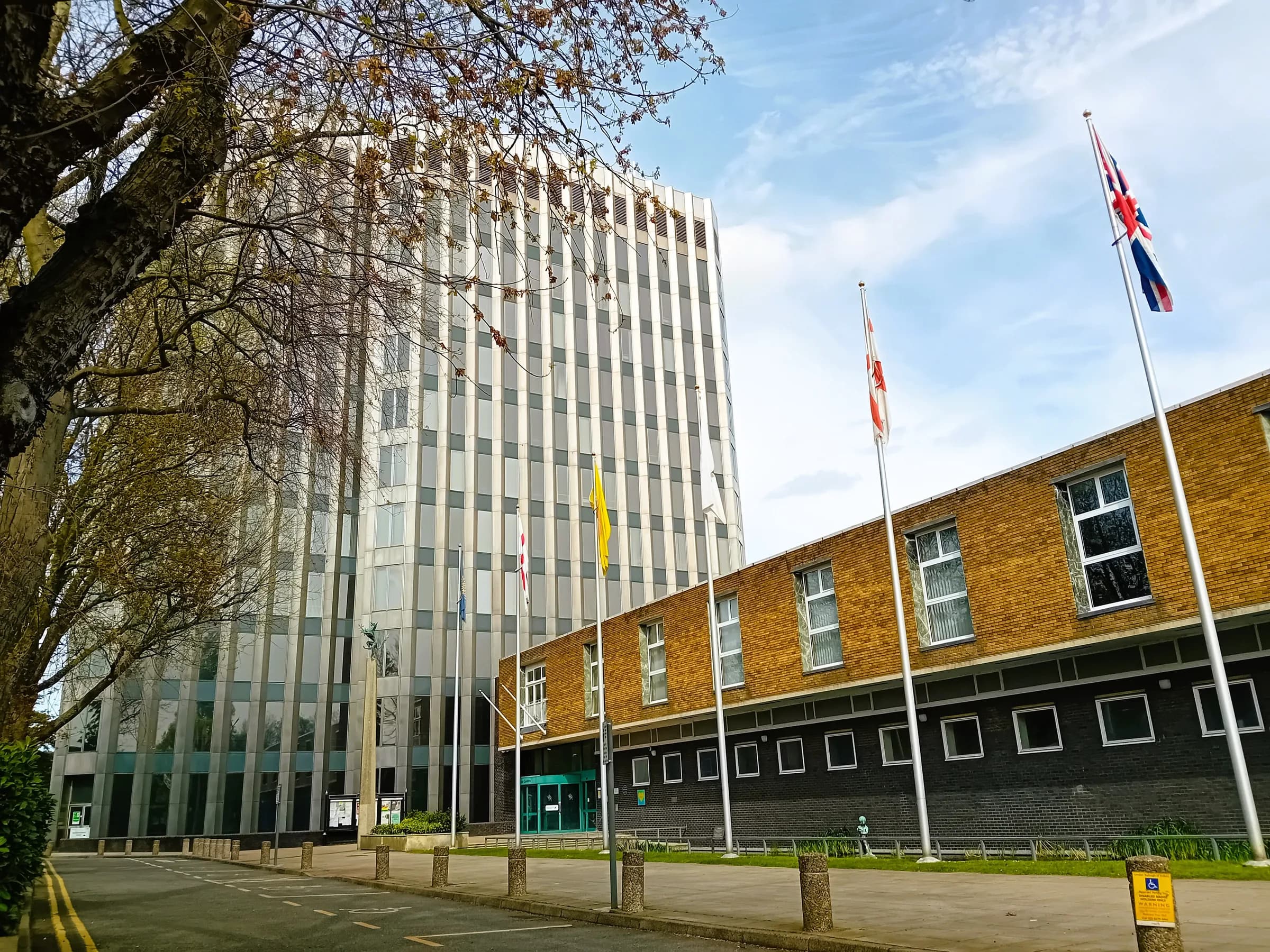 The Enfield Civic Centre council offices: a pale tower block beside a two-storey brick wing, with flags on poles and bare tree branches under a blue sky.