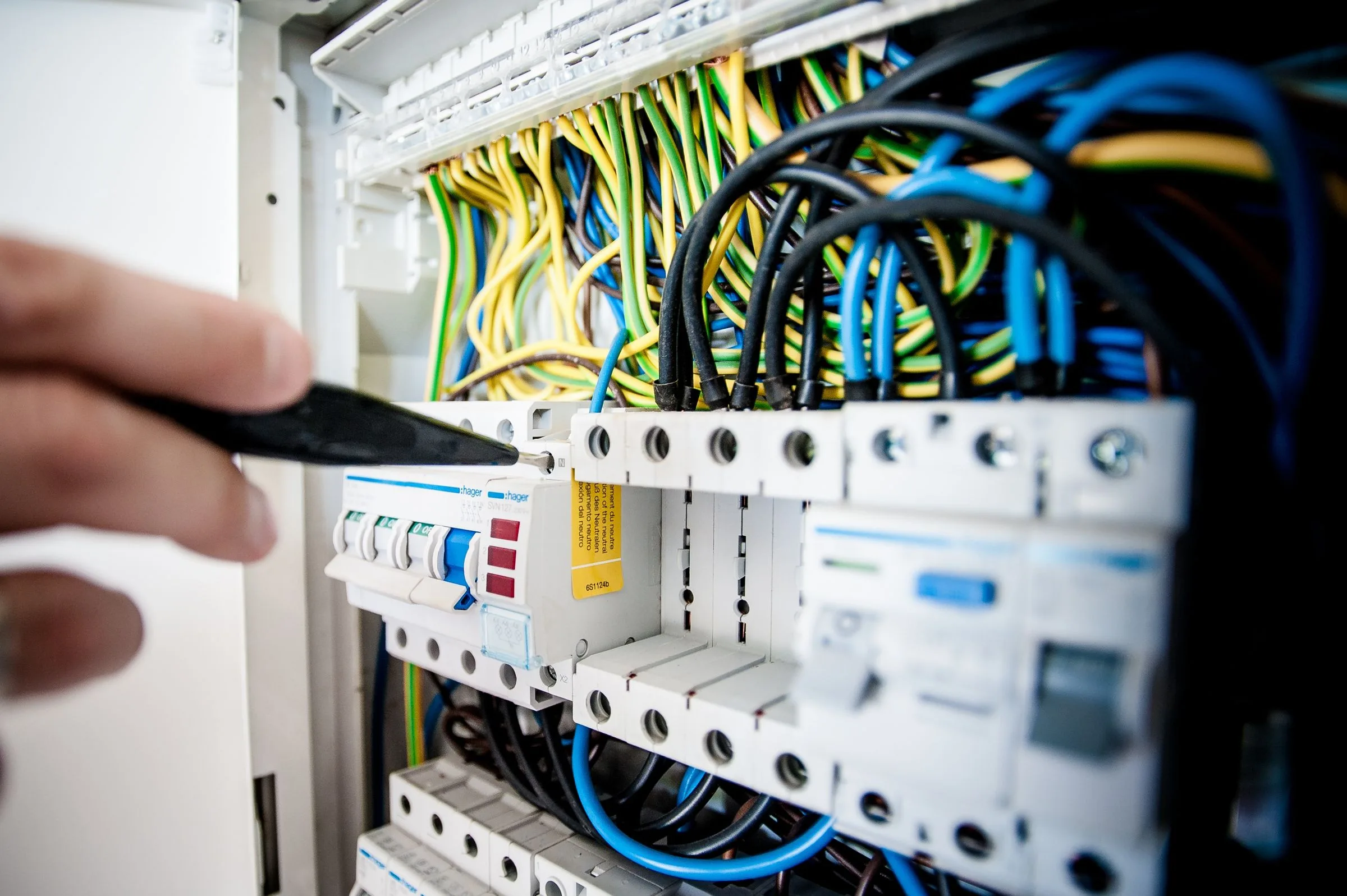 A gloved hand holding a tester probe against the circuit breakers in an open electrical distribution board full of coloured wiring.
