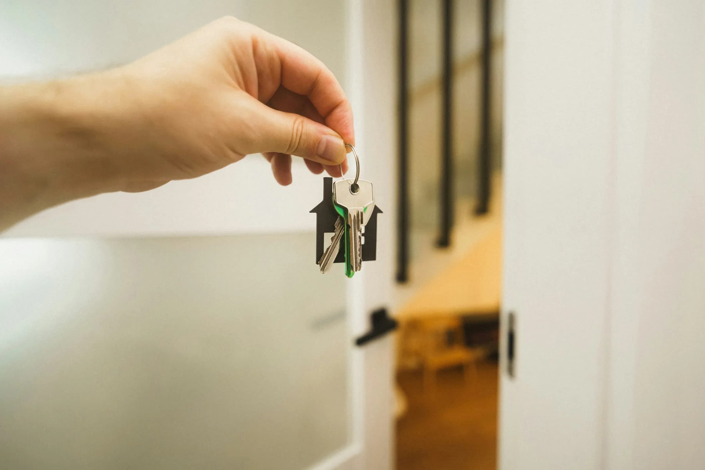 A hand holding up a set of house keys on a small house-shaped keyring in a bright hallway