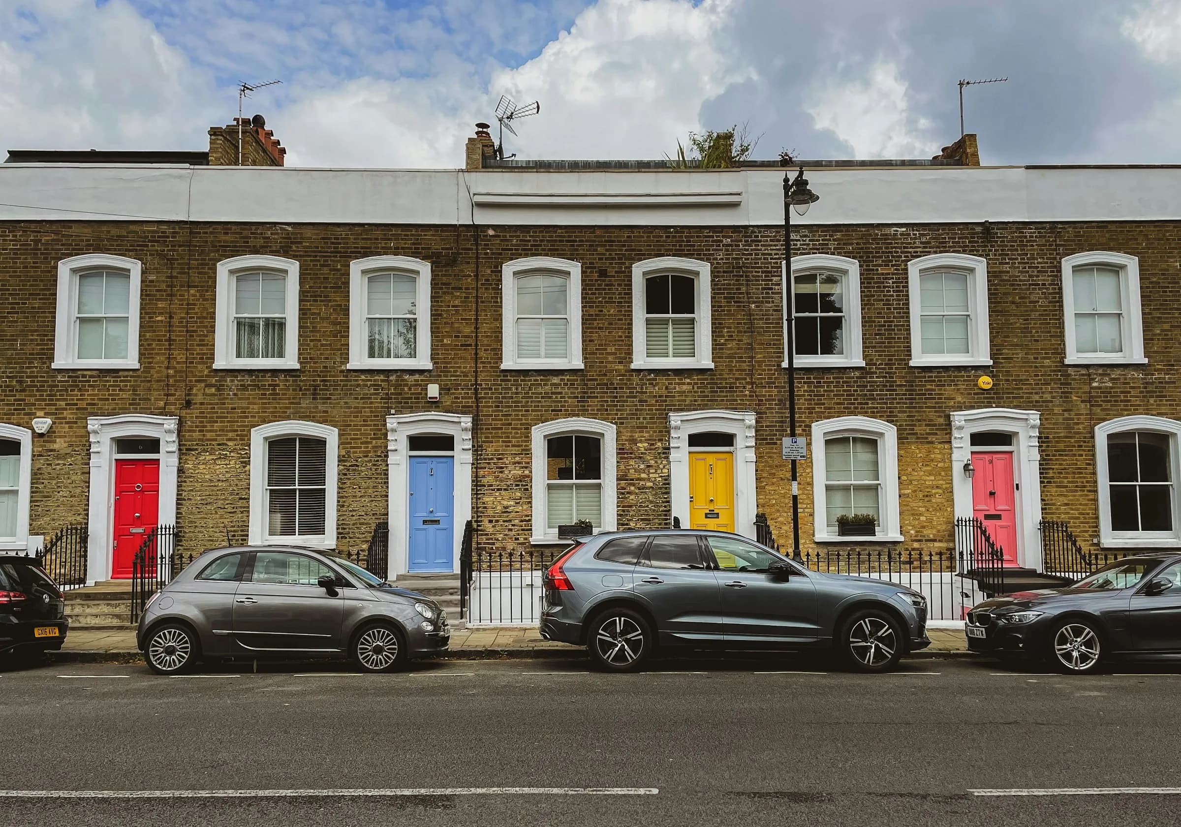 A terrace of yellow-brick Victorian houses with brightly painted front doors and cars parked along the street.