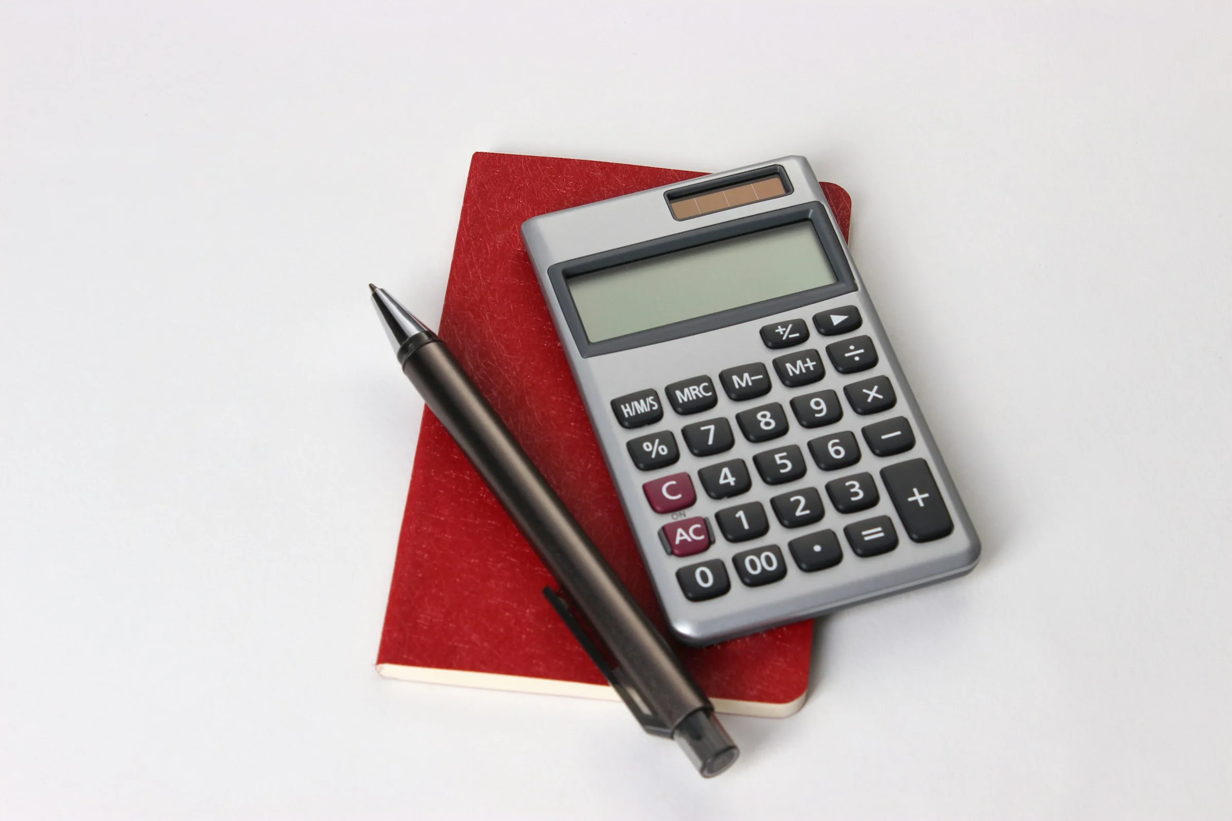 A silver pocket calculator and a black pen resting on a small red notebook on a white surface.